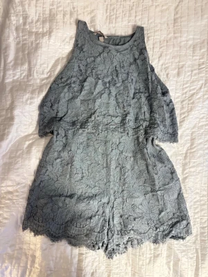 Blå/grön spets jumpsuit - Supersnygg jumpsuit med blommig spets, öppen rygg och volangdetaljer. Ärmlös modell med rund halsringning och korta ben. Storlek L men sitter som en M