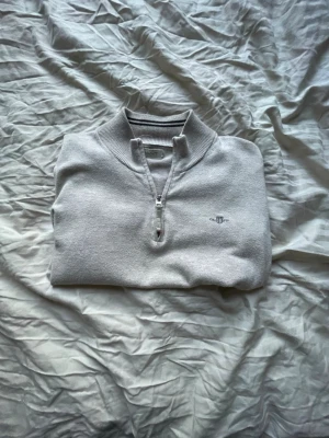 Halvzip från GANT - Viktigt “junior size Medium” Ljusgrå långärmad tröja från Gant med halv dragkedja och broderad logga på bröstet. Tröjan har ribbade muddar vid ärmslut och nederkant, samt en hög krage. Perfekt för lager-på-lager och stilren look.