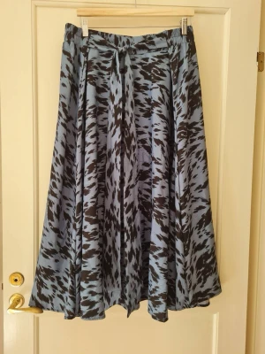 Mönstrad långkjol strl 5XL - Snygg långkjol i blått och svart med abstrakt mönster. Kjolen har resår i midjan och ett knytband framtill. Tillverkad i 100% polyester. Hela längden: 86cm. Storlek 5XL.