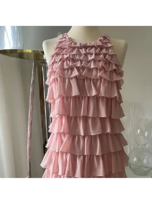 Rosa volangklänning med öppen rygg - Supersöt rosa klänning med flera lager volanger och halterneck. Klänningen har en öppen rygg med snygg detalj och är gjord i ett lätt, flowigt tyg som ger en romantisk känsla. Perfekt för dig som gillar en lekfull och feminin stil.