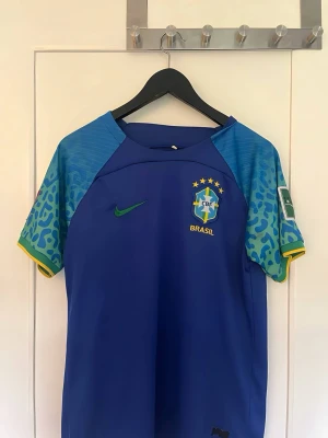 Blå Brasilien fotbollströja Nike - Snygg blå Brasilien fotbollströja från Nike med grönt Nike-märke och CBF-logga på bröstet. Ärmarna har blått och grönt mönster med gula detaljer och patch på ena ärmen. Perfekt för dig som älskar fotboll och vill visa ditt stöd för Brasilien.