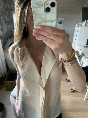 Beige ärmlös blus från VILA - Snygg beige ärmlös blus från VILA med v-ringad omlott-look och lätt transparent material. Perfekt för en stilren och fräsch outfit. Luftig och skön passform med fina detaljer framtill.