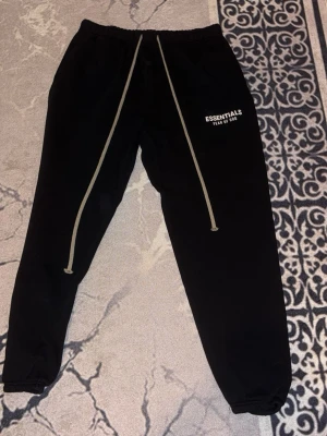 Svarta Essentials mjukisbyxor - Svarta mjukisbyxor från Fear of God Essentials med vita logotypdetaljer på vänster ben och långa beigea dragsnören. Byxorna har resår i midjan och vid bensluten för en skön och avslappnad passform. Perfekta för en chill och trendig look. Pris går att diskutera, finns även hoodie och annan färg