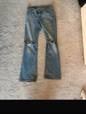 Levi's ljusblå low waist bootcut jeans - Säljer ett par ljusblå Levi's jeans med bootcut passform och slitningar vid knäna. Klassiska femficksmodellen med Levi's ikoniska läderpatch bak och röd etikett på bakfickan. Perfekta för en avslappnad och trendig look.
