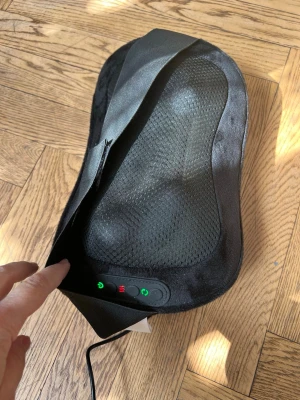 Svart elektrisk massagekudde - Elektrisk massagekudde i svart tyg med meshdetaljer och inbyggda knappar för styrning. Kudden har en ergonomisk form och ansluts med sladd och adapter. Perfekt för att massera nacke eller rygg hemma. Enkel att använda med tydliga knappar på sidan.