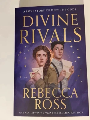 Divine Rivals av Rebecca Ross - En magisk och romantisk skönlitterär bok på engelska, 'Divine Rivals' av Rebecca Ross. Omslaget är lila med illustration av två unga personer som håller brev. Perfekt för dig som gillar fantasy, kärlek och rivalitet bland unga vuxna. Helt ny skick 