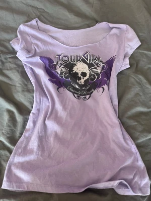 Lila t shirt med tryck - Snygg lila t-shirt  med ett stort tryck av döskalle och vingar i svart och lila på bröstet. T-shirten har korta ärmar och en feminin, figurnära passform. Perfekt för dig som gillar rockig stil.