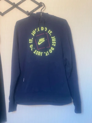 Blå Nike hoodie med neontryck - Snygg blå hoodie från Nike med neonfärgat tryck och texten 'Just Do It' i cirkel på bröstet. Klassisk huva med dragsko och stor magficka framtill. Materialet är mjukt och bekvämt, perfekt för chill eller träning.