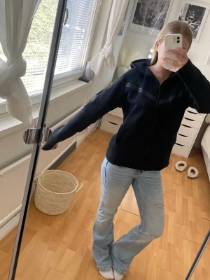 Svart hoodie med dragkedja - Svart Nike tech hoodie med dragkedja framtill och huva. Snygg detalj med mörkare band över bröstet och på ärmen. Modellen är rak och har långa ärmar. Dragkedjan är sönder och fasnar i mitten, låter dåligt men det är rätt enkelt att bara trä på sig den istället. 