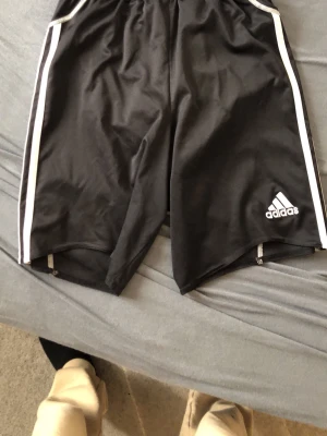 Svarta Adidas shorts med vita ränder - Svarta sportiga shorts från Adidas med klassiska vita ränder längs sidorna och Adidas-logga på ena benet. Tillverkade i ett lätt och svalt polyestermaterial, perfekta för träning eller chill. Elastisk midja för skön passform. Fick dessa shorts av en vän, vet ej hur mycket dom kostar.