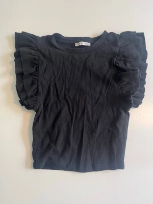 Svart topp med volangärmar från Zara - Svart ribbad topp från Zara med korta ärmar och stora volangdetaljer på axlarna. T-shirten har rund halsringning och är tillverkad i ett mjukt, stretchigt material som sitter snyggt på kroppen. Perfekt för dig som vill ha något extra till din outfit.