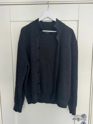 Mörkgrå ribbstickad kofta från Massimo Dutti  - Mörkgrå ribbstickad kofta från massimo Dutti med knappar framtill och lång ärm. Koftan har en klassisk rak passform och är tillverkad i ett mjukt material som passar perfekt för lager-på-lager. Snygg att matcha med jeans eller kjol.
