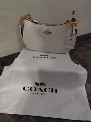 Vit axelremsväska från Coach - Snygg vit handväska från Coach med guldfärgade detaljer och logotyp framtill. Väskan har ett diskret mönster i materialet och justerbar axelrem. Perfekt för dig som vill ha en stilren och trendig accessoar från ett välkänt märke.