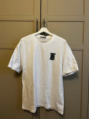 Vit t-shirt från Burberry storlek M/L - Vit t-shirt från Burberry med svart TB-logga på bröstet. Klassisk rund hals och korta ärmar. Tillverkad i mjuk bomull för en skön känsla och enkel stil.