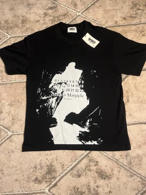 Svart t-shirt Maison Margiela - Cool svart t-shirt från Maison Margiela med stor vit abstrakt print och logga på framsidan. T-shirten har rund hals och korta ärmar. Perfekt för dig som gillar streetwear och statement-plagg.