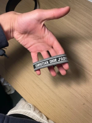 Christian Dior J'Adior armband - Snyggt vävt armband från Christian Dior med texten 'J'Adior' i vitt på svart bakgrund. Armbandet har justerbara tofsar i ändarna och en stilren, modern design som passar perfekt till din outfit.