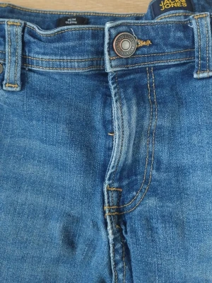 Blå slim fit jeans Jack & Jones - Slim fit jeans från Jack & Jones i klassisk blå denim med snygga slitningar och kontrastsömmar. Modellen heter Glenn och har normalhög midja, bälteshällor och knappgylf. Perfekta för en avslappnad och trendig look.