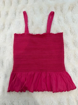 Rosa volangtopp med smock - Söt rosa topp med smala axelband och smockad överdel. Nedtill har toppen en volangkant som ger en lekfull känsla. Perfekt för dig som gillar färg och vill ha en feminin look. Materialet känns mjukt och lätt.