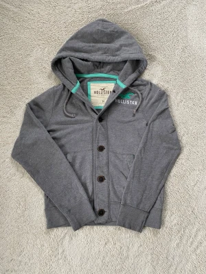 Grå hoodie från Hollister med knappar - Grå hoodie från Hollister med knäppning framtill och två fickor. Hoodien har dragsko i huvan och en liten logga på bröstet. Mjuk bomullskänsla och långärmad modell, perfekt för en chill stil.