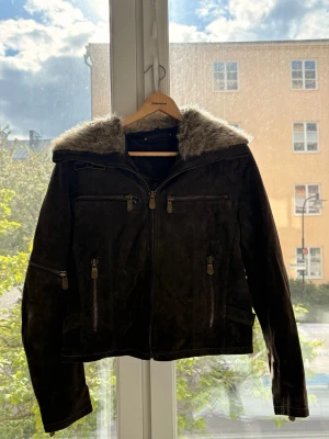 Mörkbrun skinnjacka med fuskpäls - Snygg mörkbrun skinnjacka med fuskpäls runt kragen. Jackan har flera dragkedjefickor framtill och detaljerade sömmar. Hög krage med spänne och dragkedja hela vägen upp. Perfekt för dig som gillar en cool och tidlös stil.
