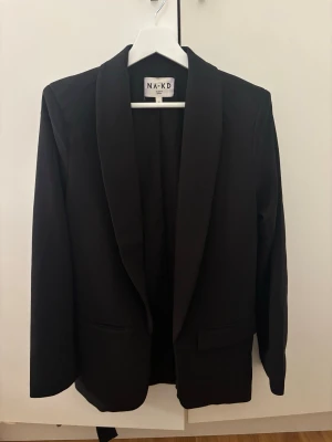 Svart kavaj från NA-KD - Stilren svart kavaj från NA-KD med klassisk krage och raka ärmar. Kavajen har två fickor framtill och en enkel, tidlös design som passar till många olika looks. Perfekt för dig som gillar en clean och modern stil.