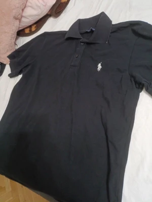 Svart pikétröja från Ralph Lauren - Klassisk svart pikétröja från Ralph Lauren med vit broderad logga på bröstet. Tröjan har krage, knappar framtill och korta ärmar. Perfekt för en stilren och enkel look. Knappen är lagad