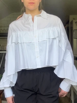 Vit volangskjorta från Zara - Säljer en vit skjorta från Zara med volangdetaljer och oversized passform. Skjortan har knappar framtill, klassisk krage och vida, långa ärmar. Perfekt för dig som gillar trendiga och unika plagg med extra volym.
