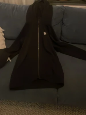 Svart Nike zip-hoodie med logga - Svart hoodie från Nike med dragkedja framtill och vit Nike-logga på bröstet. Klassisk modell med huva och dragsko, perfekt för en avslappnad streetstyle-look. Mjuk bomullskänsla och långa ärmar.