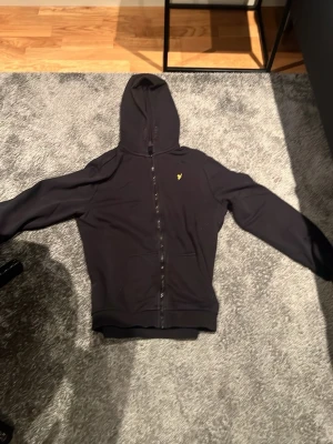 Svart hoodie med dragkedja från Lyle & Scott - Svart hoodie med dragkedja från Lyle & Scott. Klassisk modell med huva, fickor framtill och gul logga på bröstet. Perfekt för en avslappnad stil. Mjuk bomullsblandning och ribbade muddar vid ärmslut och nederkant.