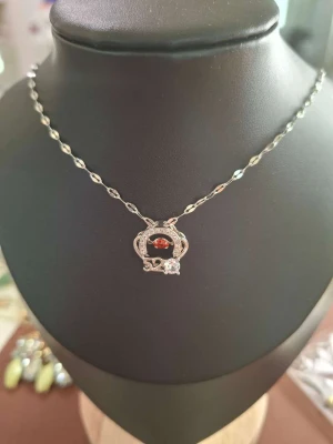 Silverhalsband med röd sten - Ett elegant halsband i silverfärgad metall med en tvinnad kedja och ett hänge med en röd pärla i mitten. Hänget har en rund form med dekorativa detaljer och en klar sten nedtill. Perfekt för att lyfta din outfit.
