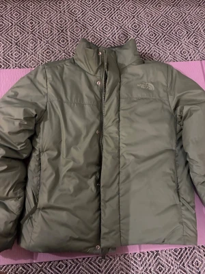 Olivgrön pufferjacka från The North Face - Snygg olivgrön pufferjacka från The North Face med hög krage och dragkedja framtill. Jackan har diskret logga på bröstet och är tillverkad i ett slitstarkt, vattenavvisande material. Perfekt för kalla dagar när du vill hålla stilen och värmen.