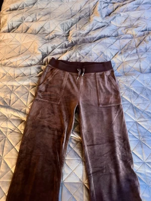 Bruna Juicy couture mjukisbyxor  med fickor - Mysiga bruna juicy couture mjukisbyxor i velour med bred resår och dnören i midjan. Byxorna har två stora framfickor och två bakfickor med knappdetaljer. Perfekta för en chill och avslappnad stil.