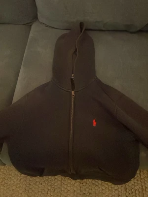 Mörkblå hoodie från Polo Ralph Lauren - Mörkblå hoodie med dragkedja från Polo Ralph Lauren. Klassisk design med röd broderad logga på bröstet och huva. Perfekt för en avslappnad och stilren look. Mjuk bomullskänsla och bekväm passform.