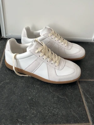 Maison Margiela Gats - Tjena! Säljer ett par Gats från Maison Margiela med beige mockadetaljer och ljusa skosnören. Skorna har en klassisk låg design och en brun gummisula. Perfekta för dig som gillar stilrena och trendiga sneakers.  Dessa är oanvända        Storlek 44. Nypris 6000kr❌ mitt pris 1799✅
