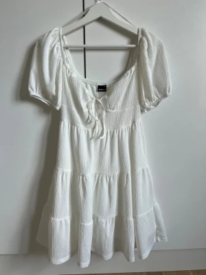 Vit kortklänning från Gina Tricot - Superfin vit kortklänning från Gina Tricot med puffärmar, knytning vid bysten och volangdetaljer. Klänningen är tillverkad i ett lätt, strukturerat tyg som ger en luftig känsla. Perfekt för varma dagar och enkel att styla med sneakers eller sandaler. Perfekt student eller midsommar klänning. Klänningen har endast använts en gång, så är i väldigt bra skick!
