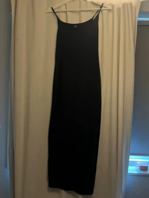 Svart långklänning med smala axelband - En stilren marinblå långklänning med smala axelband och rak passform. Klänningen har en minimalistisk design och är tillverkad i ett mjukt, slätt material som ger en elegant känsla. Perfekt för dig som gillar enkel och tidlös stil.