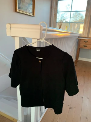 Svart kortärmad kofta från Gina Tricot - Stilren svart kofta från Gina Tricot med korta ärmar och rund halsringning. Koftan har en enkel knäppning framtill med en knapp och är tillverkad i ett mjukt material som passar perfekt till lager-på-lager-stil.