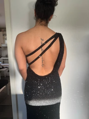 Svart glittrig långklänning oneshoulder - Svart långklänning med glittriga paljetter och snygg öppen rygg. Klänningen har oneshoulder-design med två dekorativa band över ryggen. Perfekt för dig som vill sticka ut med en elegant och modern look. Bara använts en gång i 2-4 timmar. Nypris: 6500kr