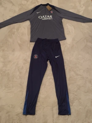 PSG tracksuit - Snyggt träningsset från Paris Saint-Germain med långärmad grå tröja och mörkblå byxor. Tröjan har PSG-logga, Nike-logga och Qatar Airways-tryck. Byxorna har PSG-märke och Nike-logga. Pris kan diskuteras. 