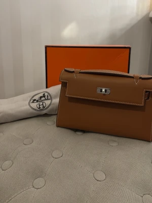 Brun  kuvertväska i läder - Säljer en brun  kuvertväska i slätt läder med silverfärgat vridlås och vita kontrastsömmar. Klassisk och stilren design med tillhörande dustbag och box. Perfekt accessoar för dig som gillar lyxiga detaljer.