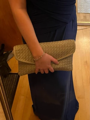 Ljusgrå flätad kuvertväska - Snygg ljusgrå kuvertväska med flätad design. Väskan har ett klassiskt kuvertformat lock och stängs med magnetknapp. Perfekt storlek för det viktigaste och enkel att matcha till olika outfits. Har ett band som går att knäppa på och av beroende på om man vill använda den som handväska