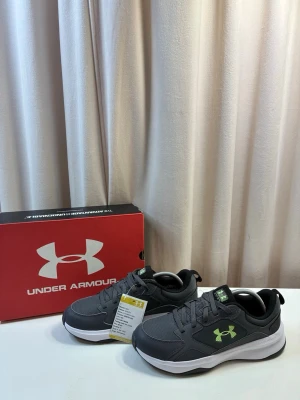 Under Armour sneakers - Snygga grå sneakers från Under Armour med limegrön logga på sidan. Skorna har en chunky vit sula och ovandel i mesh och syntetmaterial för bra andningsförmåga. Perfekta för dig som gillar sportig stil och vill ha bekväma skor till vardags.