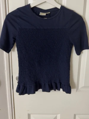 Mörkblå topp med smock från Gina Tricot - Säljer en mörkblå topp från Gina Tricot med smockad framsida och volangkant nedtill. Toppen har korta ärmar och rund halsringning, perfekt för dig som gillar en enkel men ändå söt stil.