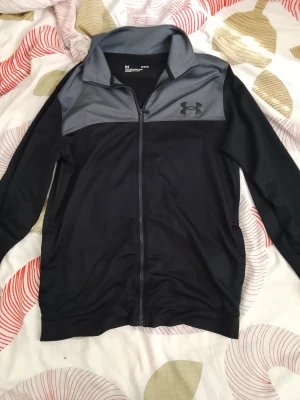 Svart och grå zip-hoodie från Under Armour - Snygg svart och grå zip-hoodie från Under Armour med dragkedja hela vägen och hög krage. Loggan syns på bröstet och materialet är mjukt och sportigt, perfekt för en aktiv stil. Långa ärmar och stilren design.
