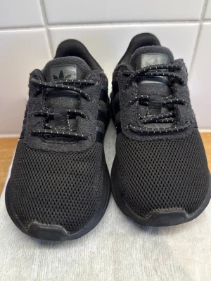  Adidas sneakers storlek 21 - Snygga helsvarta sneakers från Adidas med klassiska tre ränder på sidan. Ovandelen är i mesh och mockaliknande material, med svarta skosnören och Adidas-logga på plös och häl. Perfekta för en stilren och sportig look.