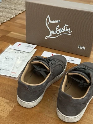 Gråa Christian Louboutin sneakers - Snygga gråa sneakers från Christian Louboutin i mocka med klassisk låg siluett. Skorna har vita sulor och beige innersida, samt matchande grå skosnören. Perfekta för dig som gillar exklusiva och stilrena sneaker, På skornas knappar har två lossnat men syns knappast!