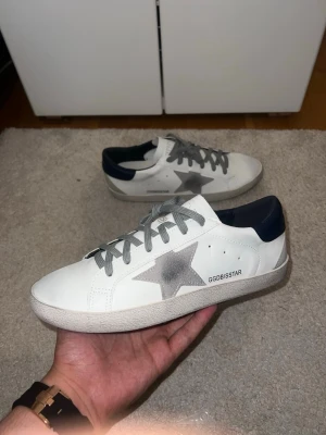 Golden Goose sneakers - Golden Goose sneakers i vitt läder med grå stjärndetalj på sidan, grå skosnören och mörkblå hälkappa. Klassisk låg modell med rund tå och platt sula. Perfekta för en stilren och trendig look.