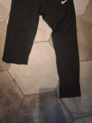 Svarta träningsbyxor från Nike - Svarta träningsbyxor från Nike med smal passform och diskret vit logga på höften. Byxorna är tillverkade i ett stretchigt material som passar perfekt för sport och träning. Klassisk design med långa ben och bekväm midja.