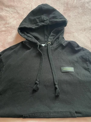 Svart hoodie från COLLUSION - Snygg svart hoodie från COLLUSION med dragsko i huvan och logga på bröstet. Tillverkad i tungt bomullsmaterial och har en klassisk känguruficka framtill. Perfekt för en chill och avslappnad stil. Unisex storlek M