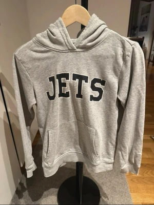 Grå hoodie med JETS-tryck - Snygg grå hoodie med svart JETS-tryck på bröstet. Klassisk känguruficka framtill och skön huva. Tillverkad i mjukt sweatshirtmaterial, perfekt för en avslappnad och sportig stil. Texten något urtvättad men annars i bra skick 